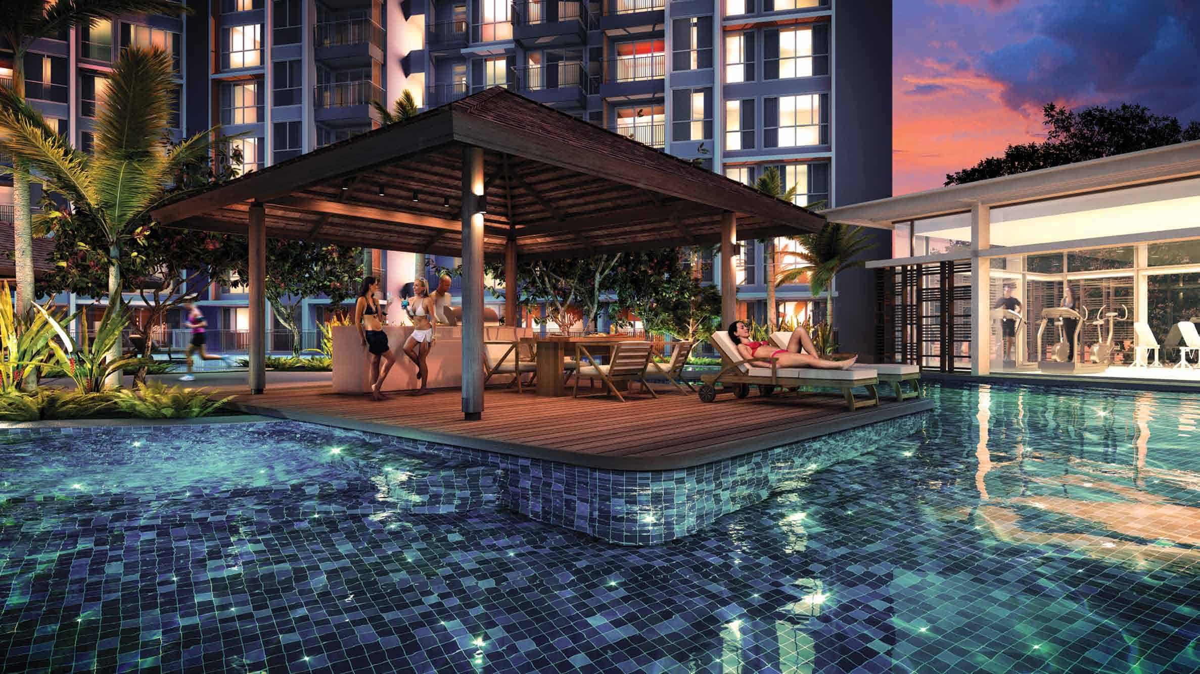 Evening view of the poolside pavilion at Qbay Residences, featuring residents lounging and socializing under a wooden gazebo surrounded by lush landscaping, with a well-lit gym visible through glass windows in the background.